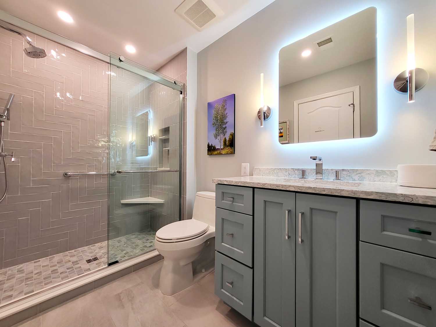 Modern bathroom with a glass-enclosed shower, blue vanity with marble countertop, backlit mirror, wall-mounted lights, and a toilet. A small painting hangs on the wall.
