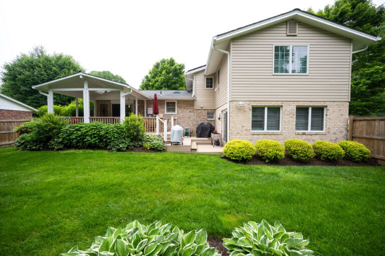 A suburban backyard with a well-kept lawn, a covered patio, grill, shrubs, and a tan, two-story house with white trim and fenced perimeter.