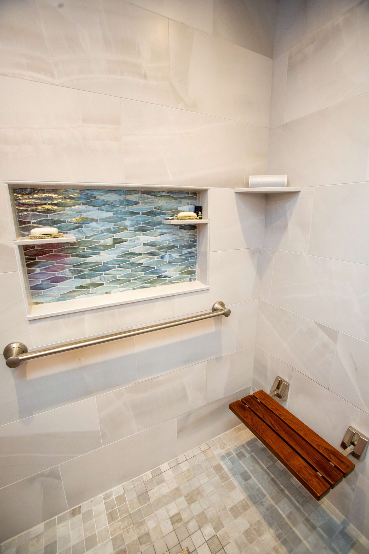Accessible shower with a grab bar, fold-down wooden bench, tiled niche with shelves, and light-colored wall and floor tiles.