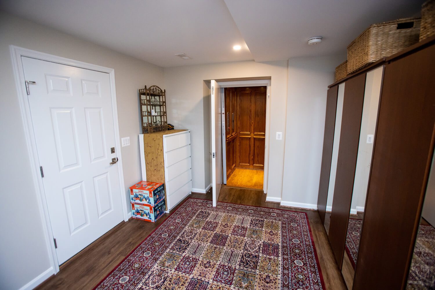 A room with a patterned rug, a white door, a dresser, stacking toy blocks, a mirror, dark wardrobes, and an open door revealing a wood-paneled elevator.