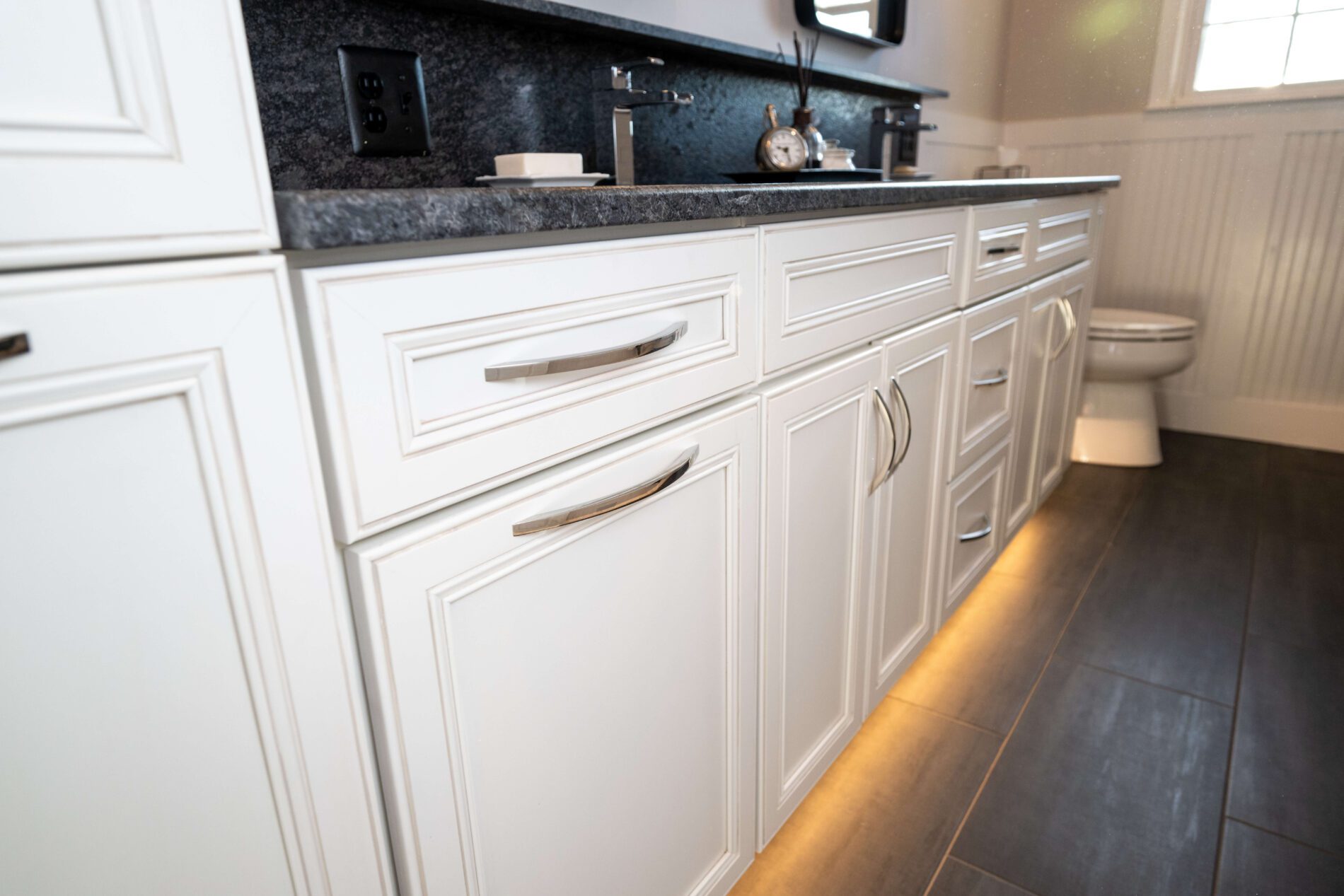 White bathroom cabinets with silver handles, dark countertop, and under-cabinet lighting; a toilet is visible in the background on the right.