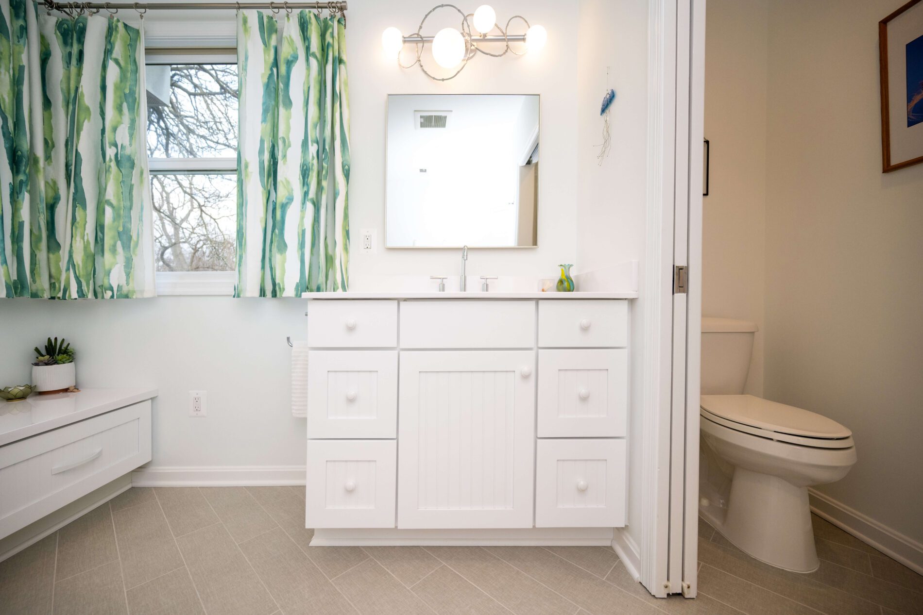 A bathroom with a white vanity, mirror, green patterned curtains, a toilet, and light-colored tiled floor.