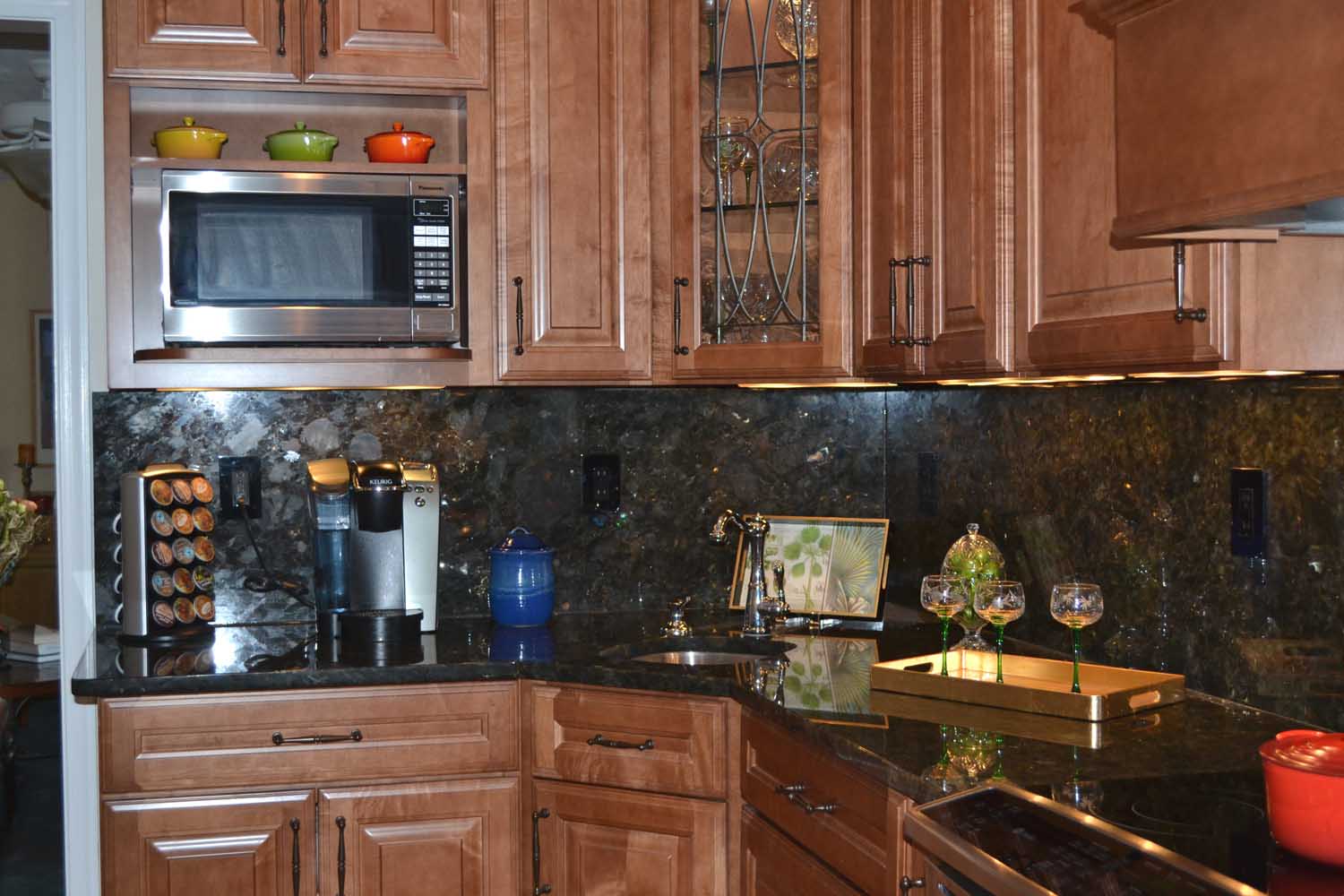 Modern kitchen with wood cabinets, black marble countertops, a microwave, coffee maker, wine glasses on a tray, and colorful pots on display above the microwave.