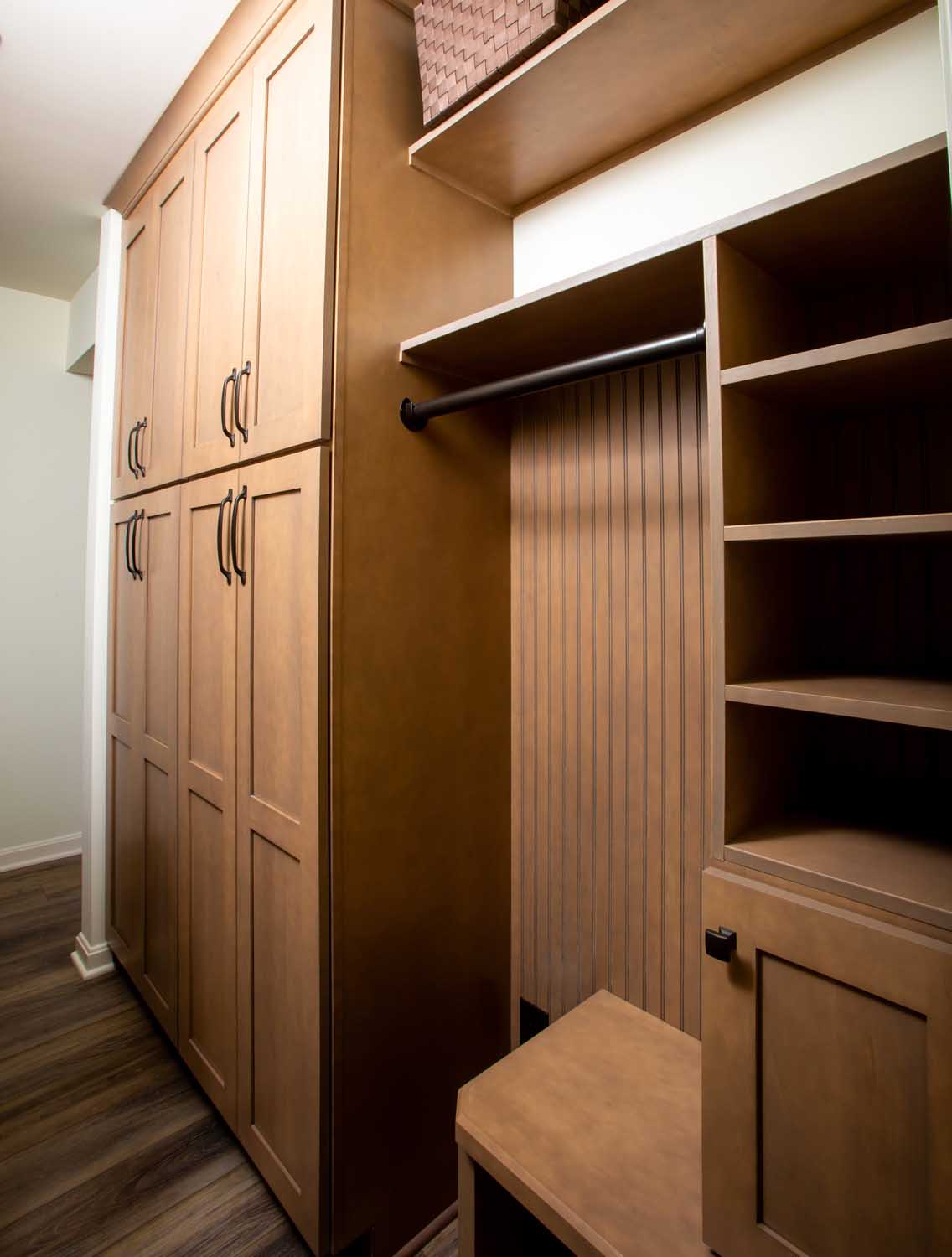 Custom wooden closet with cabinets, shelves, a hanging rod, and a bench, set against a wall with wood flooring.