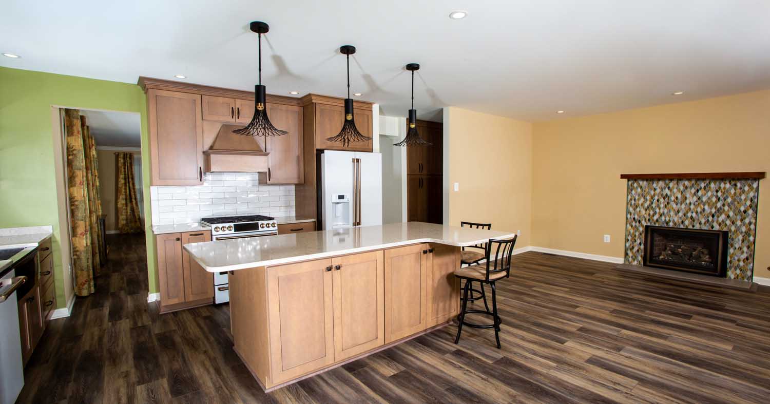 Modern kitchen with wooden cabinets, an island with barstool, pendant lights, stainless appliances, and an open living area with fireplace and patterned tile.