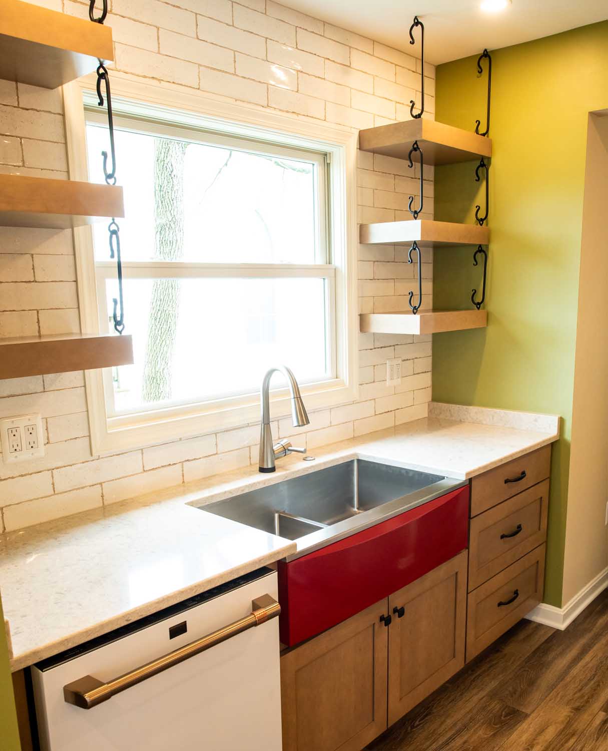 Modern kitchen with a large window, farmhouse sink with a red front, wooden cabinets, open shelves with black brackets, and white tile backsplash.