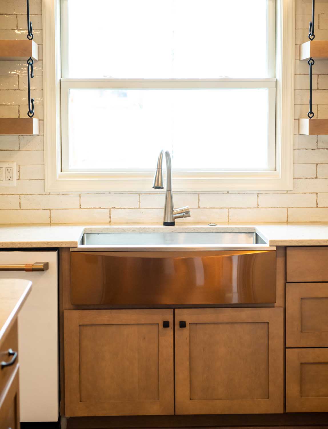 A modern kitchen sink with a stainless steel faucet is set in a wooden cabinet beneath a large window, with white tiled walls and open shelves on either side.