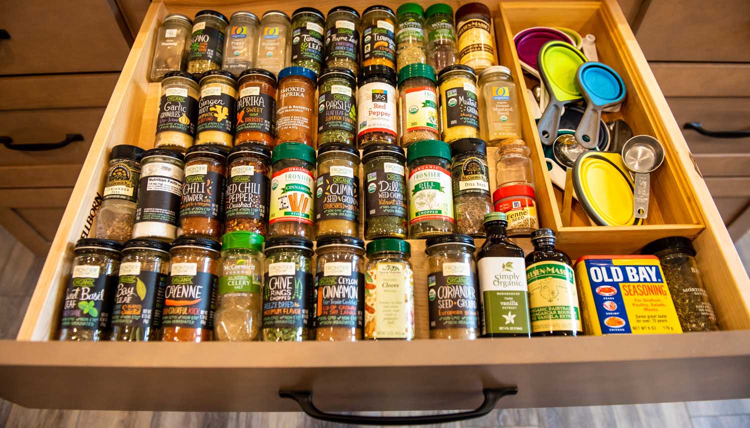 A kitchen drawer neatly organized with jars of spices, a box of Old Bay seasoning, measuring spoons, and other small kitchen tools.