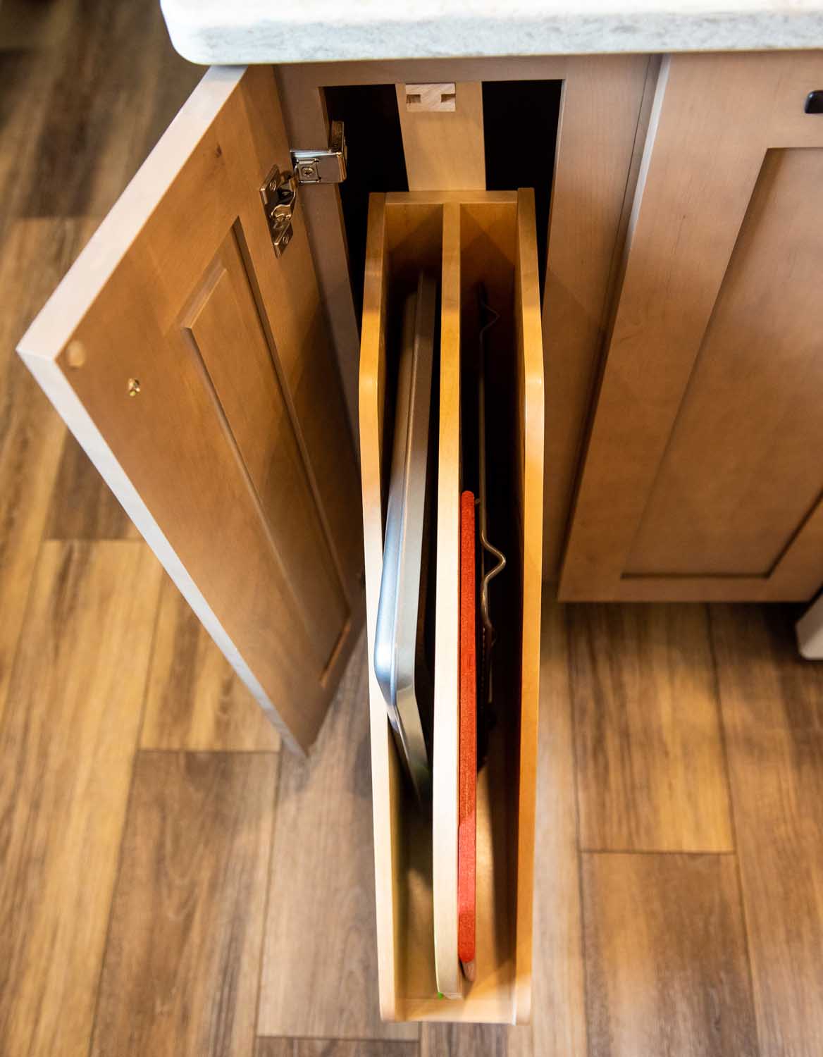 A wooden kitchen cabinet with a vertical pull-out rack containing baking trays and cutting boards, partially opened on a wood floor.