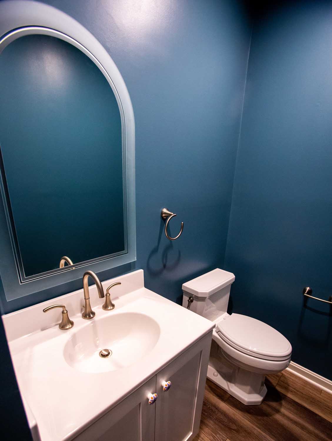 A small bathroom with blue walls, a white vanity and sink, an arched mirror, a toilet, and wood-look flooring.