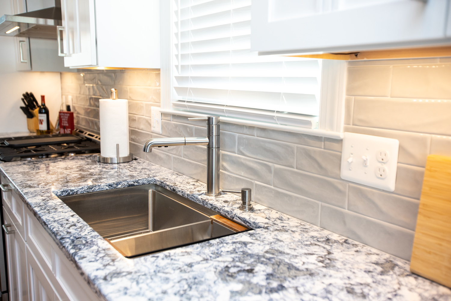 Modern kitchen with a marble countertop, stainless steel sink, soap dispenser, paper towel holder, and stove; light gray subway tile backsplash and window with blinds above the sink.