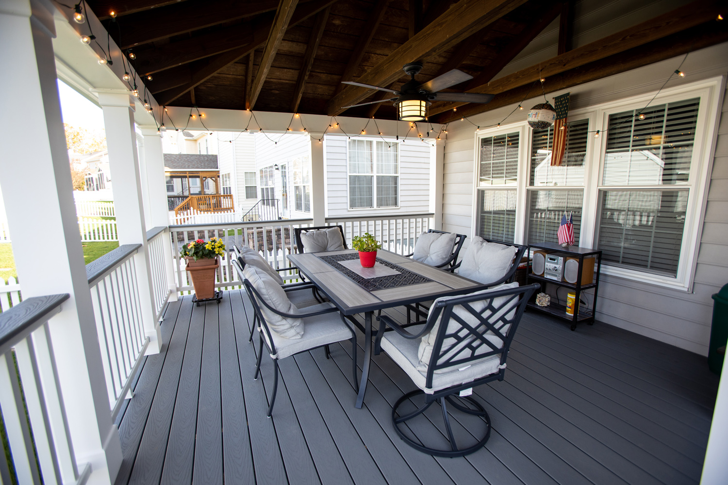 Covered porch with string lights, a table and six cushioned chairs, potted plants, and a ceiling fan; house and yard visible in background.