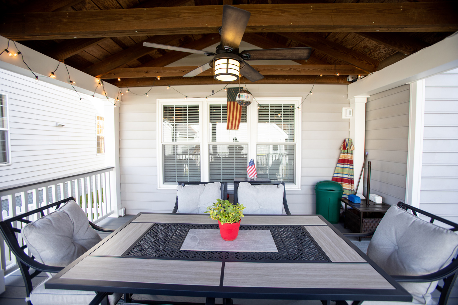 Covered patio with a ceiling fan, string lights, cushioned chairs around a table, a small plant centerpiece, and an American flag hanging in front of a window.