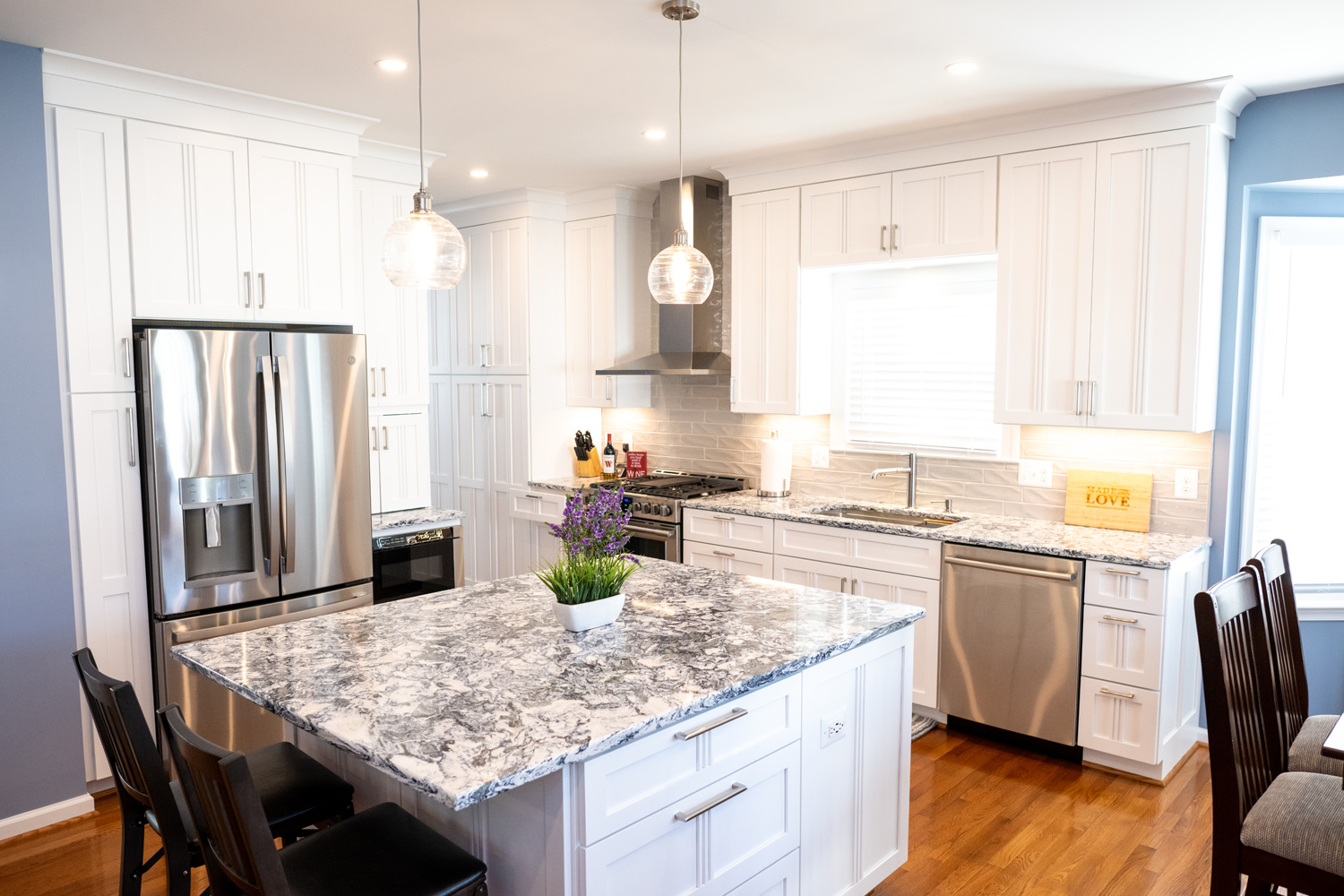 Modern kitchen with white cabinets, stainless steel appliances, marble island countertop, and a potted plant centerpiece. Dining chairs line one side of the island.