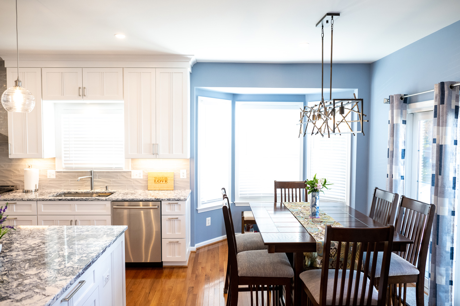 Modern kitchen and dining area with white cabinets, stainless steel appliances, granite countertops, blue walls, and a wooden dining set near a bay window with natural light.