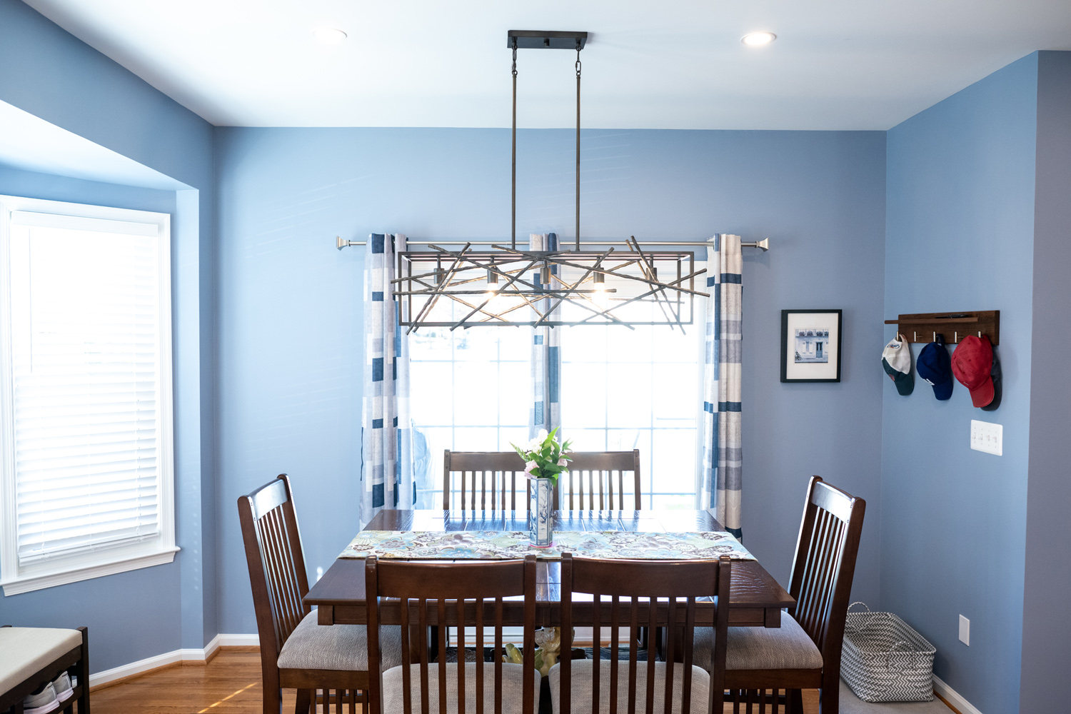 A dining room with a wooden table and six chairs, blue walls, a window with blue curtains, and hats hanging on a wall rack. A modern light fixture hangs above the table.