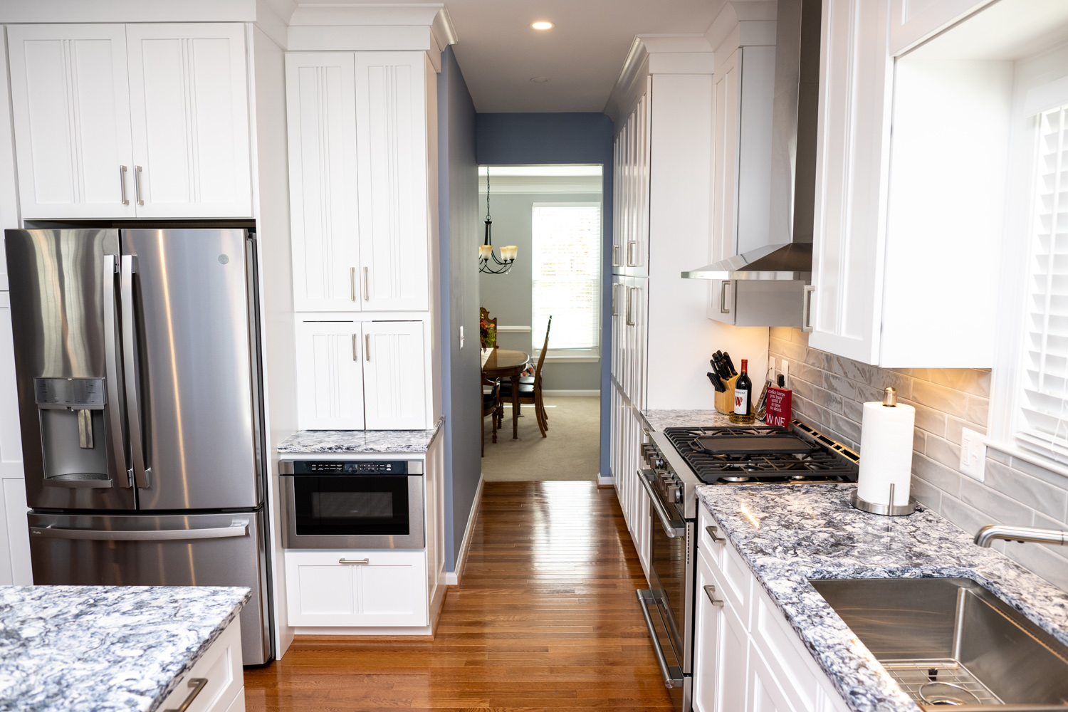 Modern kitchen with white cabinets, stainless steel appliances, marble countertops, and hardwood floors; dining room visible through hallway in the background.