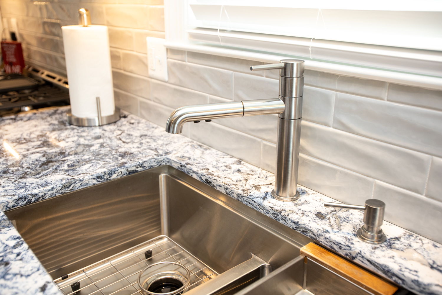Modern stainless steel kitchen faucet and sink with a marble countertop, gray tile backsplash, and a paper towel holder in the background.