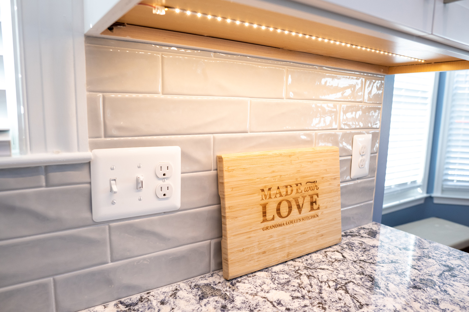 A kitchen countertop with a cutting board that reads "Made with Love" leaning against a light gray tiled backsplash, featuring two electrical outlets.