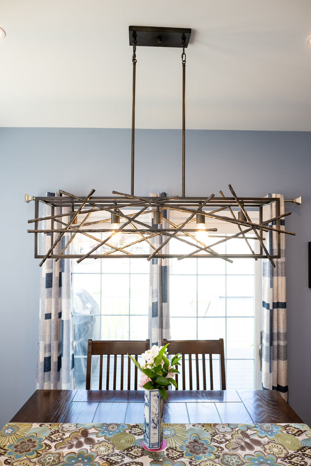 A modern chandelier with a geometric metal frame hangs above a wooden dining table with a floral vase, set in front of large windows with blue curtains.
