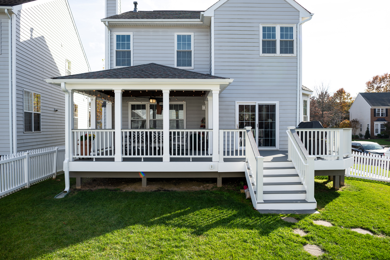 A two-story gray house with white trim features a covered back porch, white railings, stairs leading down to a fenced yard, and a grill on the deck area.
