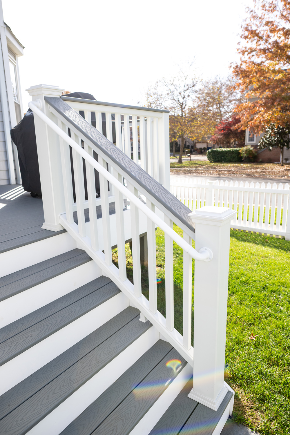 Outdoor staircase with gray steps and white railings, leading to a grassy yard with a white picket fence and trees with autumn foliage in the background.