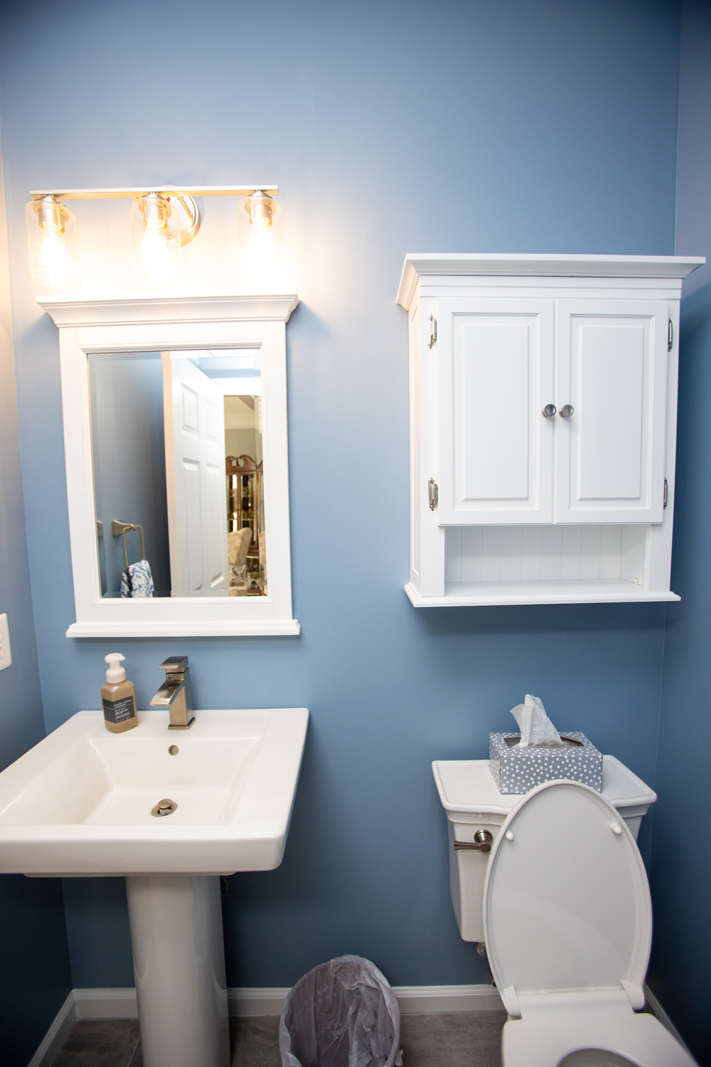 A bathroom with blue walls, a white pedestal sink, a mirror with lights above, a wall cabinet, and a toilet with a tissue box on top.