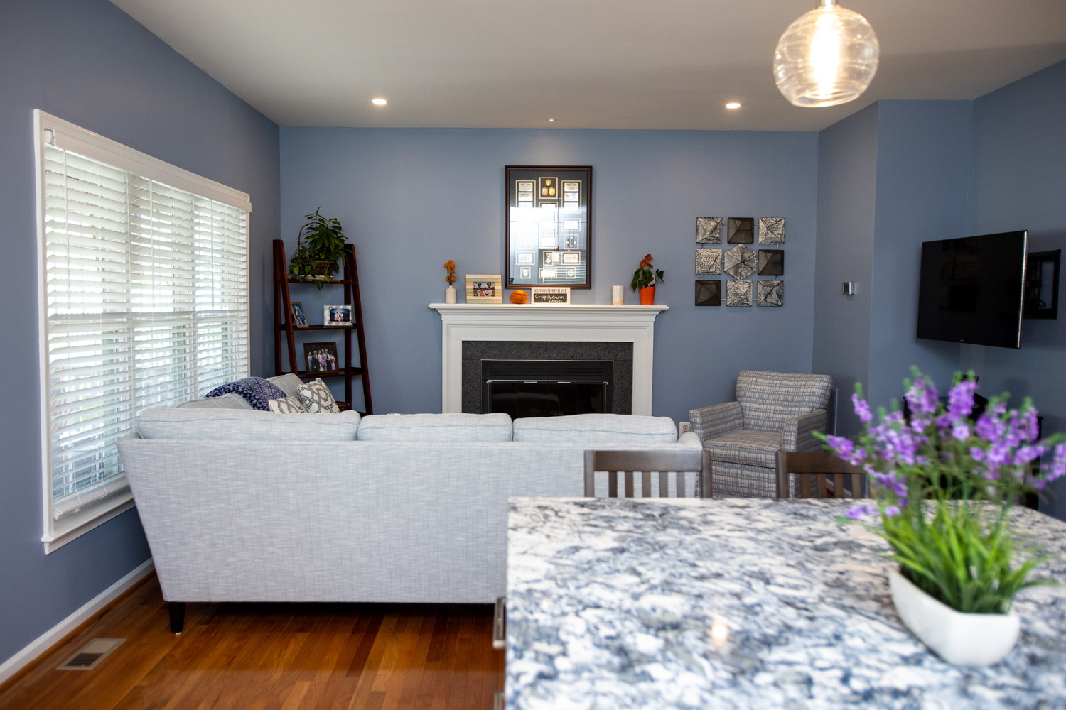 A modern living room with blue walls, a light gray sofa, a fireplace, a wall-mounted TV, and a marble countertop with a potted plant in the foreground.