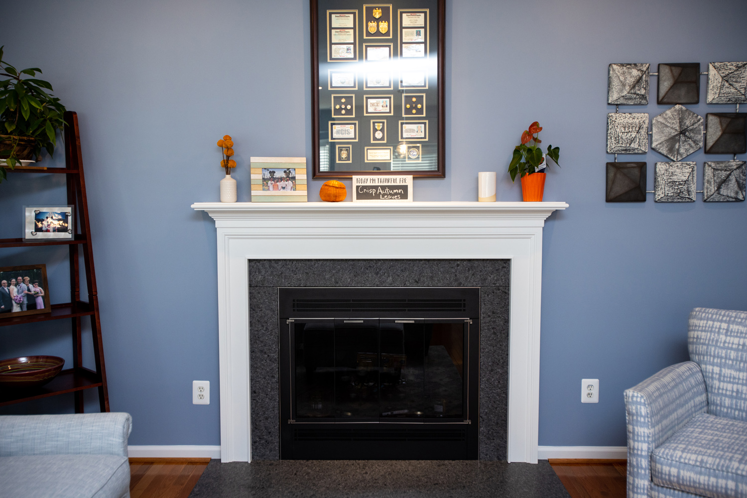 A white fireplace with a black insert is centered on a blue wall, decorated with framed memorabilia, plants, and small decorative items. Blue furniture and a wooden floor are visible.
