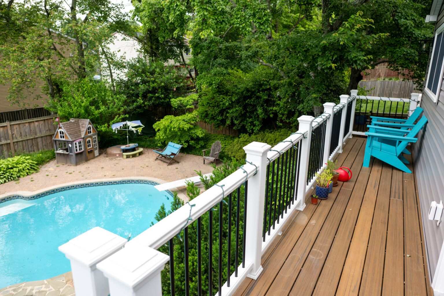 A backyard with a swimming pool, a small playhouse, patio seating, umbrella, and greenery, viewed from a wooden deck with blue chairs and potted plants.
