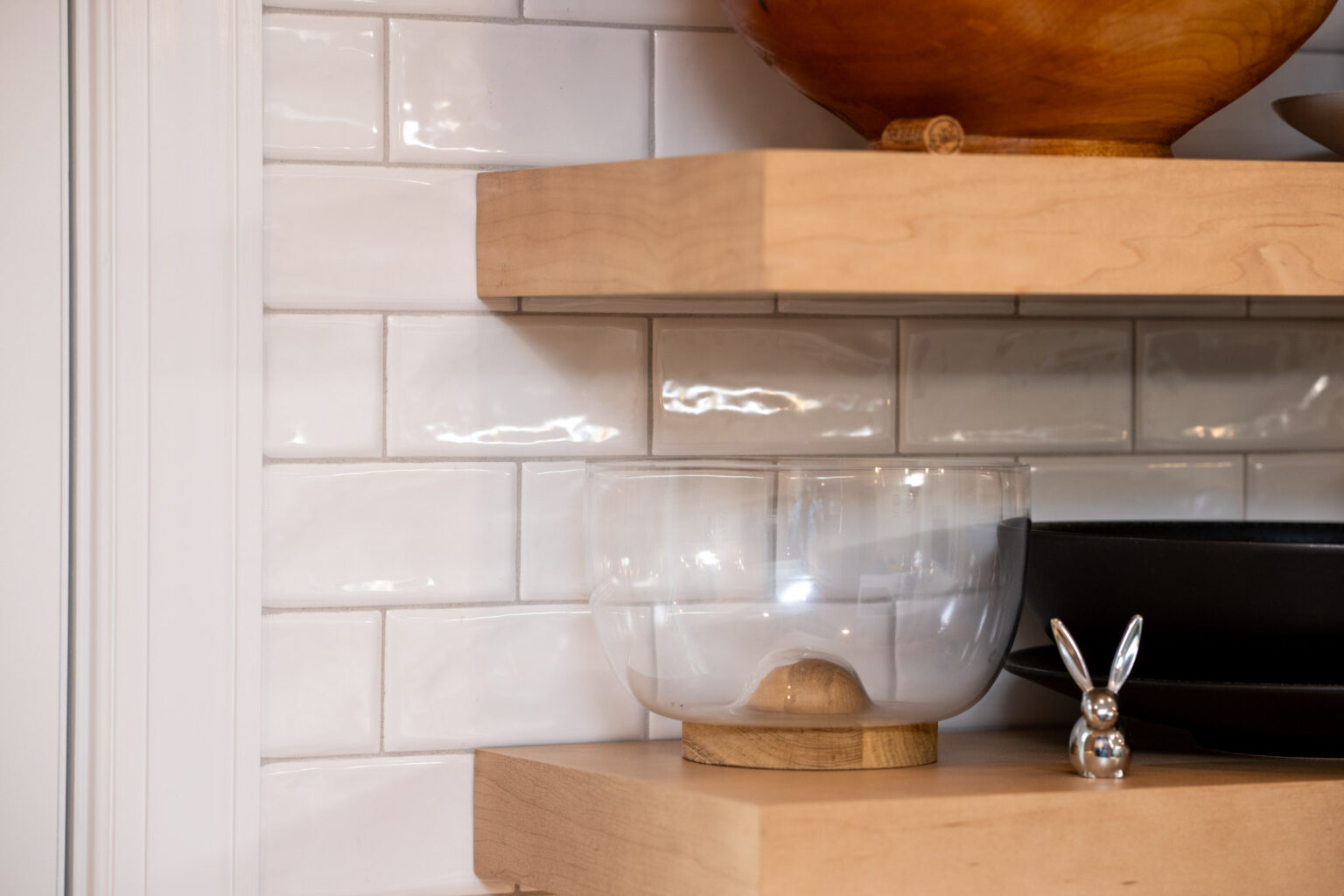 A clear glass bowl with a wooden base, a black dish, and a silver rabbit figurine on light wooden shelves against a white subway tile backsplash.