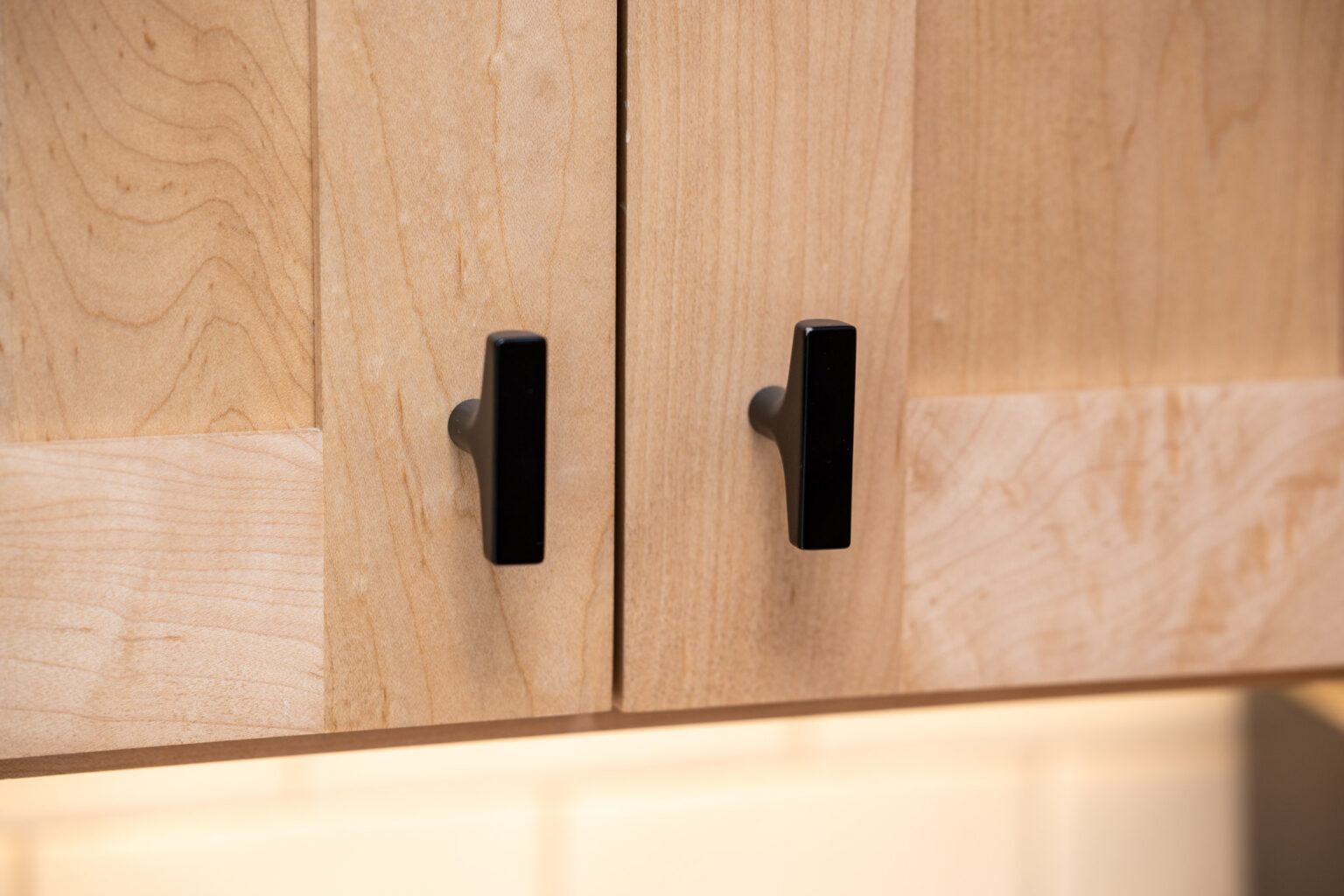 Close-up of two light wood cabinet doors with black rectangular handles, set above a beige tile backsplash.