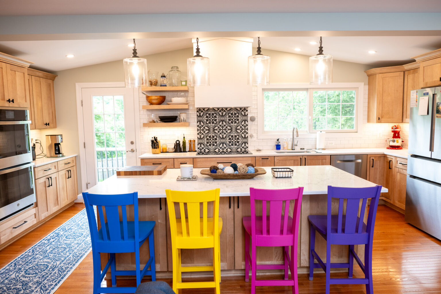 Modern kitchen with wooden cabinets, a central island, and four colorful chairs in blue, yellow, and magenta. Pendant lights hang overhead; large windows provide natural light.