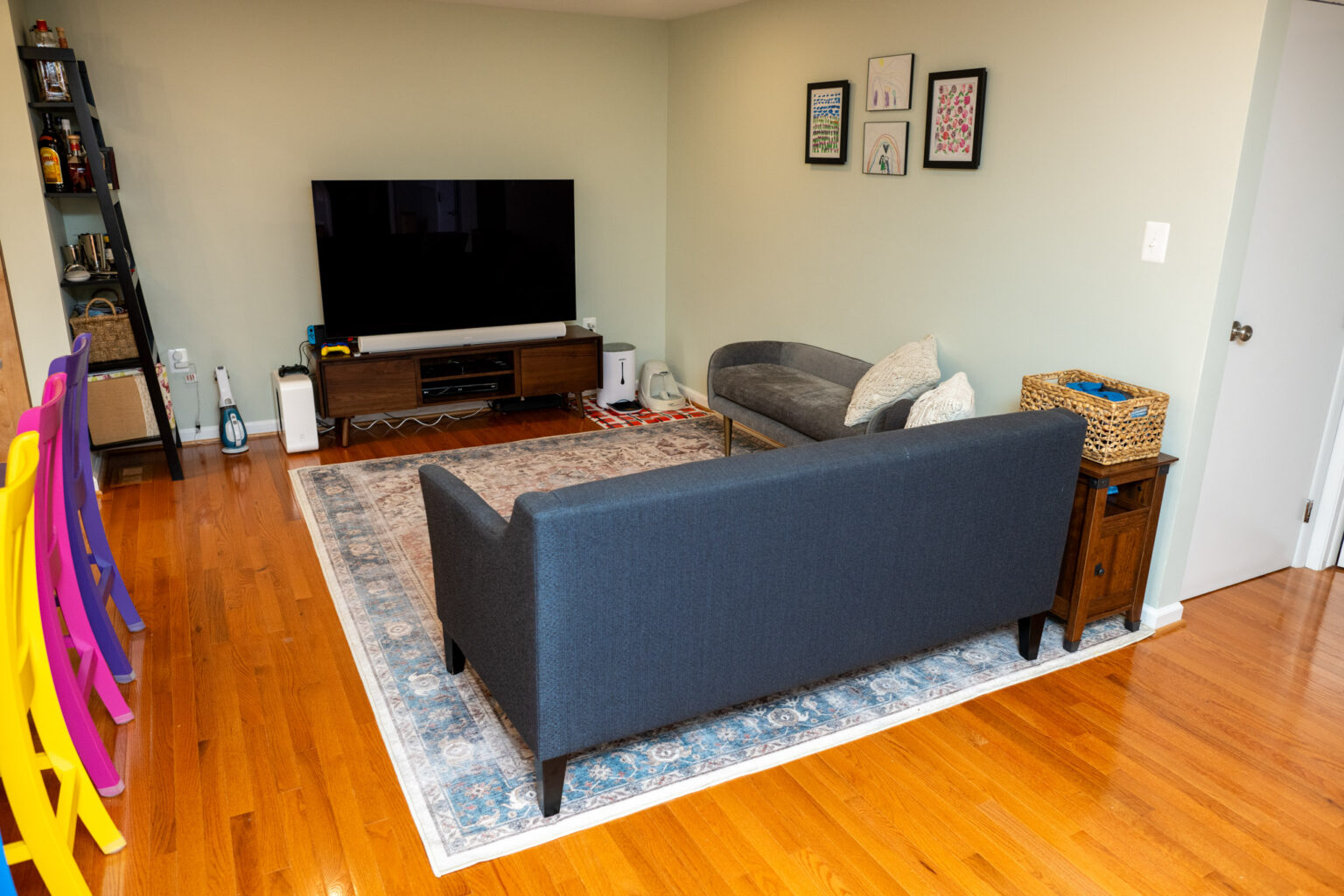 A living room with a dark sofa, a TV on a stand, a rug on wooden floors, colorful chairs on the left, and framed pictures on a light-colored wall.