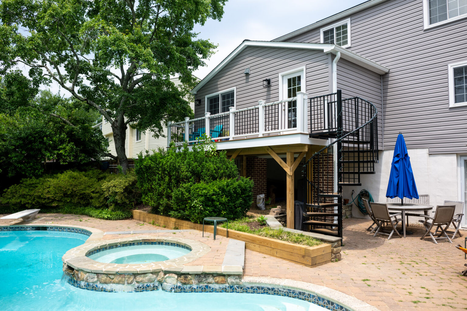 Backyard with an in-ground pool, attached hot tub, wooden deck with spiral staircase, and patio furniture near a gray house.