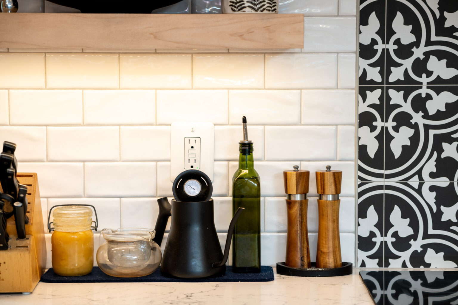 A kitchen countertop with a knife block, glass jars, an oil dispenser, an olive oil bottle, and salt and pepper mills against white tile and patterned black tile backsplash.