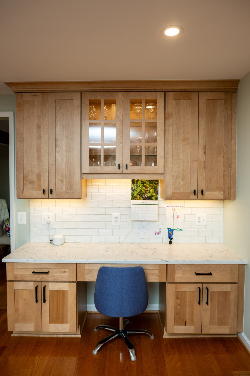 A built-in wooden desk with upper and lower cabinets, a blue office chair, under-cabinet lighting, and a white subway tile backsplash.
