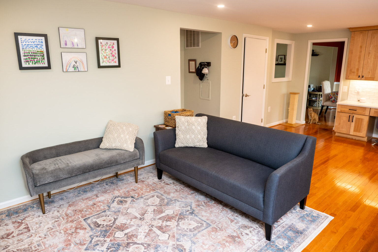 A living room with a gray loveseat, a gray bench, two throw pillows, framed art on the wall, a patterned rug, and hardwood floors. A kitchen is visible in the background.