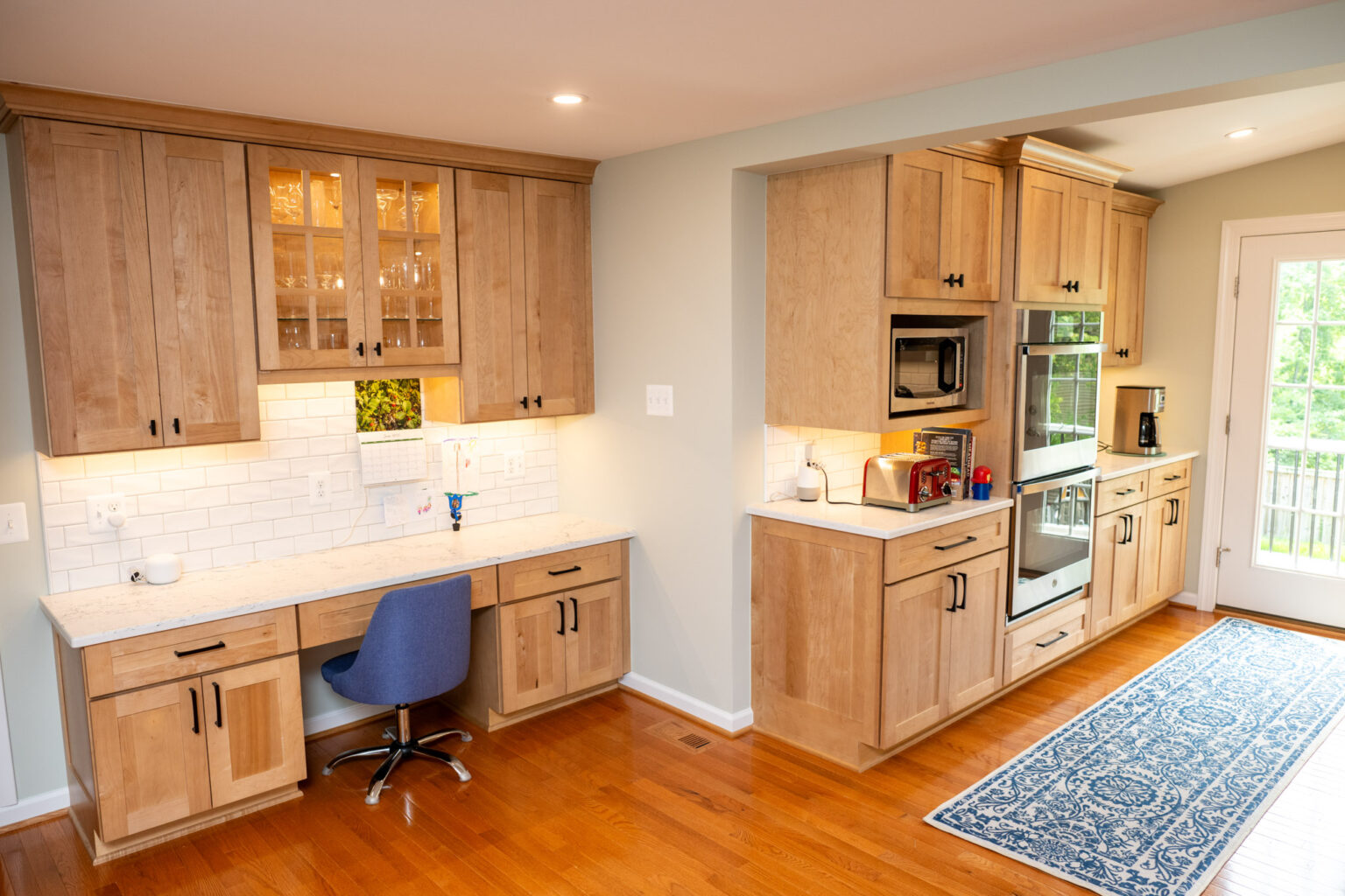 A modern kitchen with light wood cabinets, white countertops, built-in appliances, a small desk area with a blue chair, and a door leading outside.