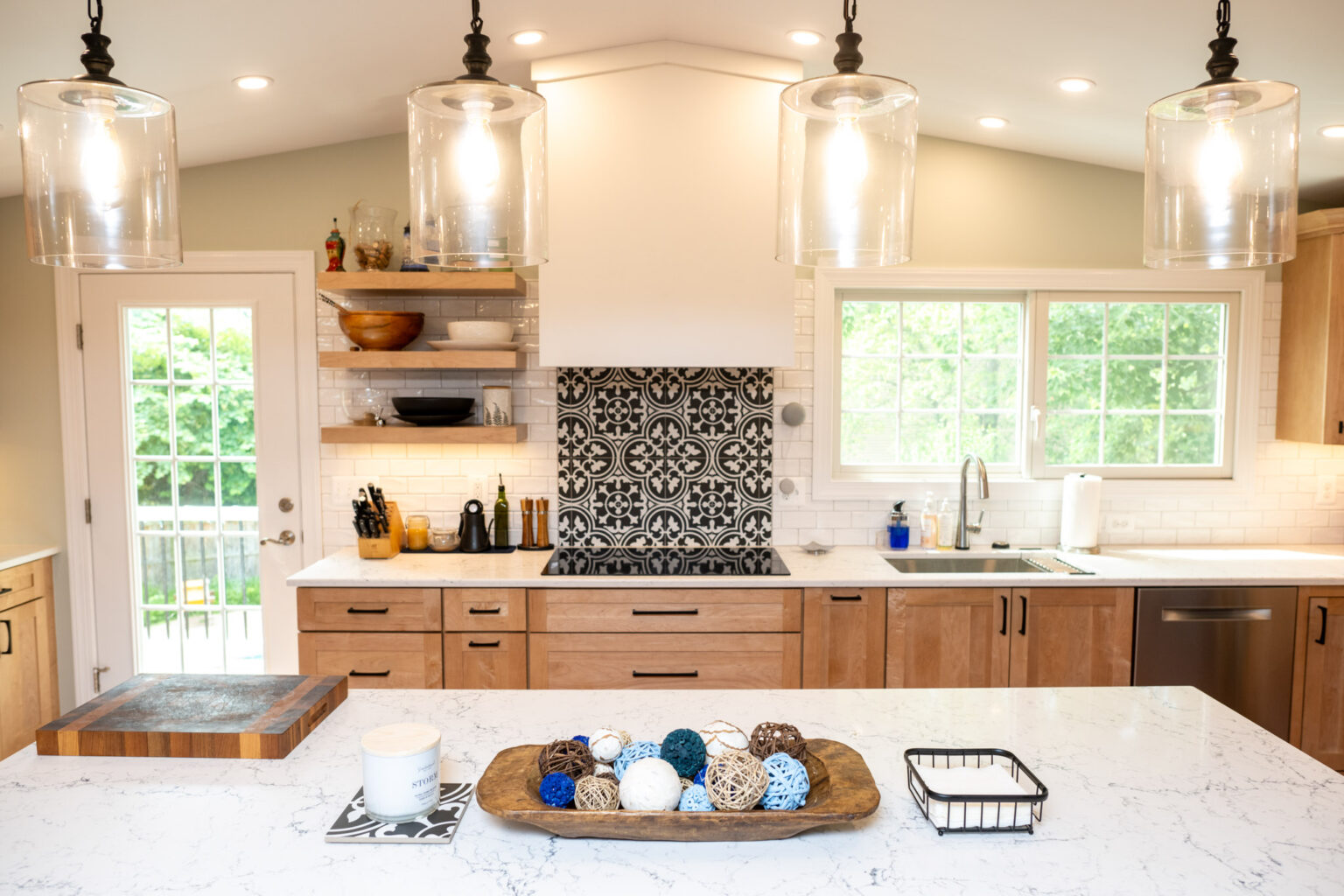 Modern kitchen with wood cabinets, white countertops, three pendant lights, a patterned backsplash, open shelves, and a tray with decorative balls on the island.