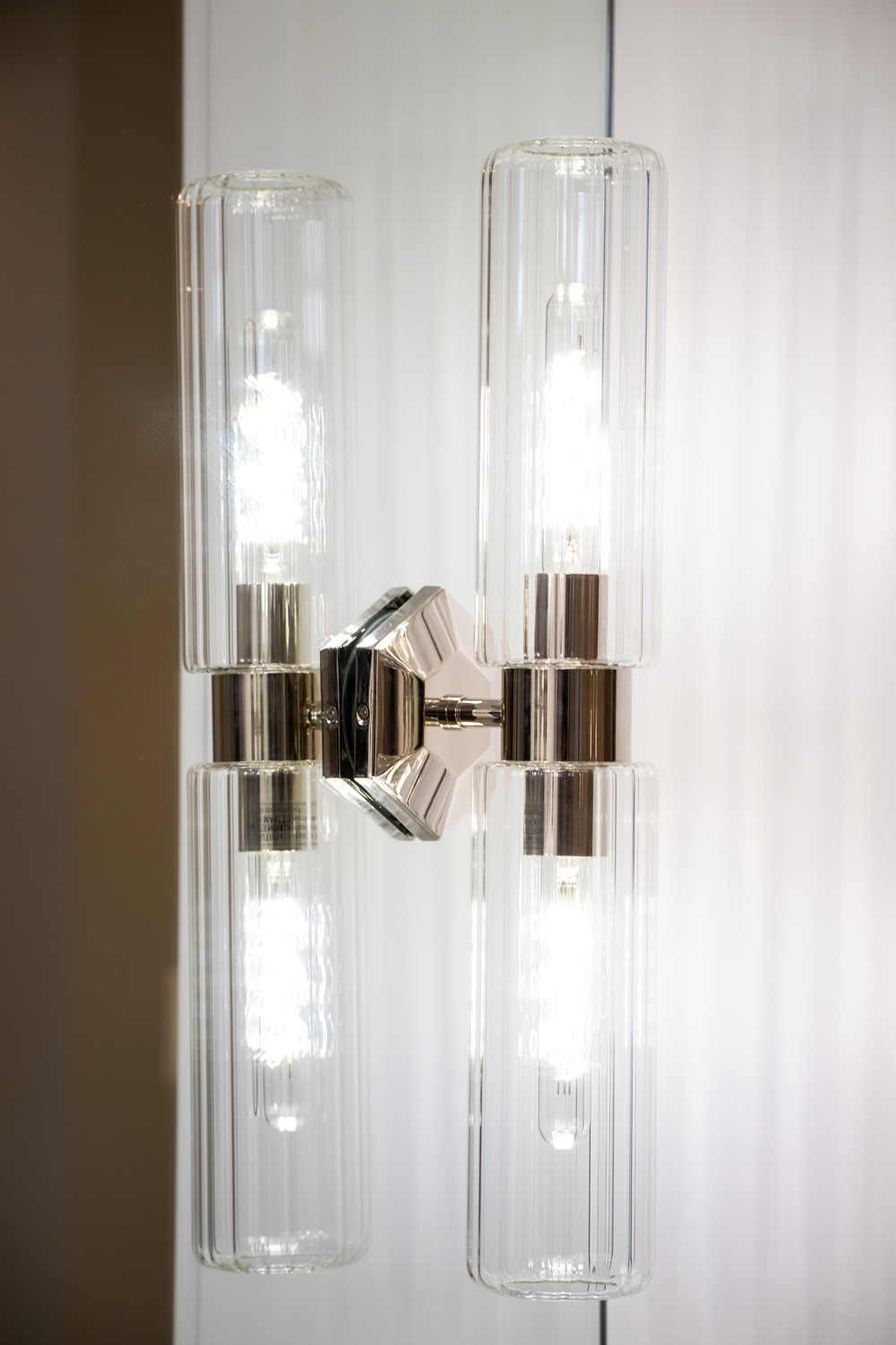 A modern wall sconce with two vertical LED bulbs encased in clear ribbed glass covers, mounted on a metallic base against a reflective surface.