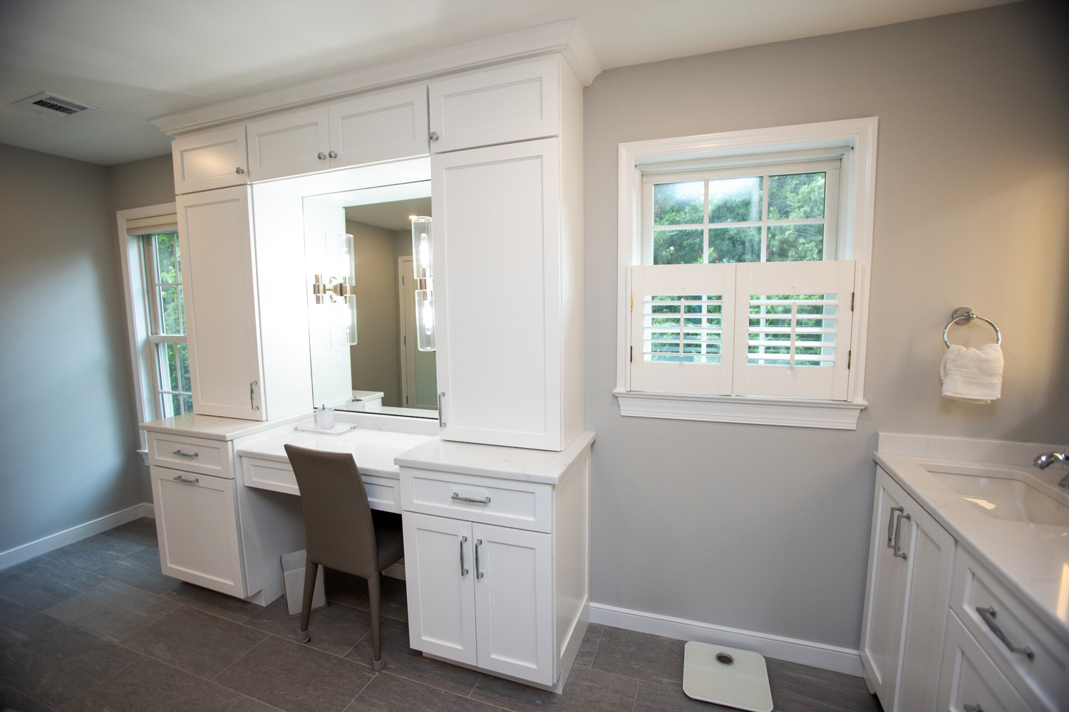 A modern bathroom with white cabinetry, a vanity with a mirror and chair, a window with shutters, a towel ring, and a digital scale on the tiled floor.