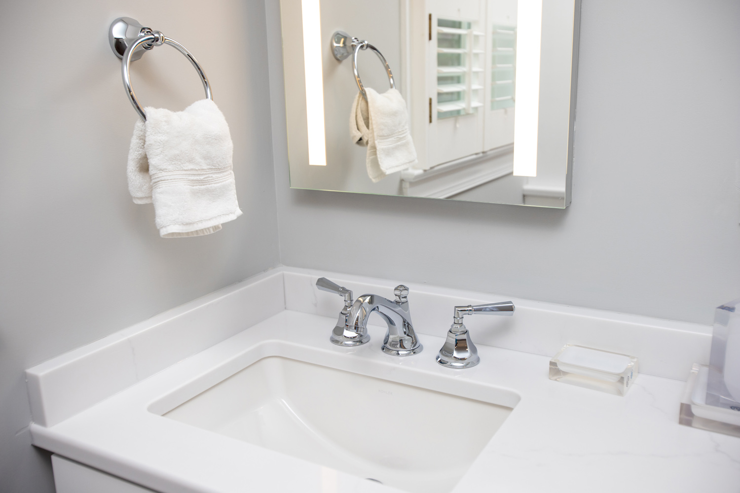 Modern bathroom sink with chrome faucet, a rectangular mirror, white countertop, folded hand towel on a ring, and a soap dish on the counter.