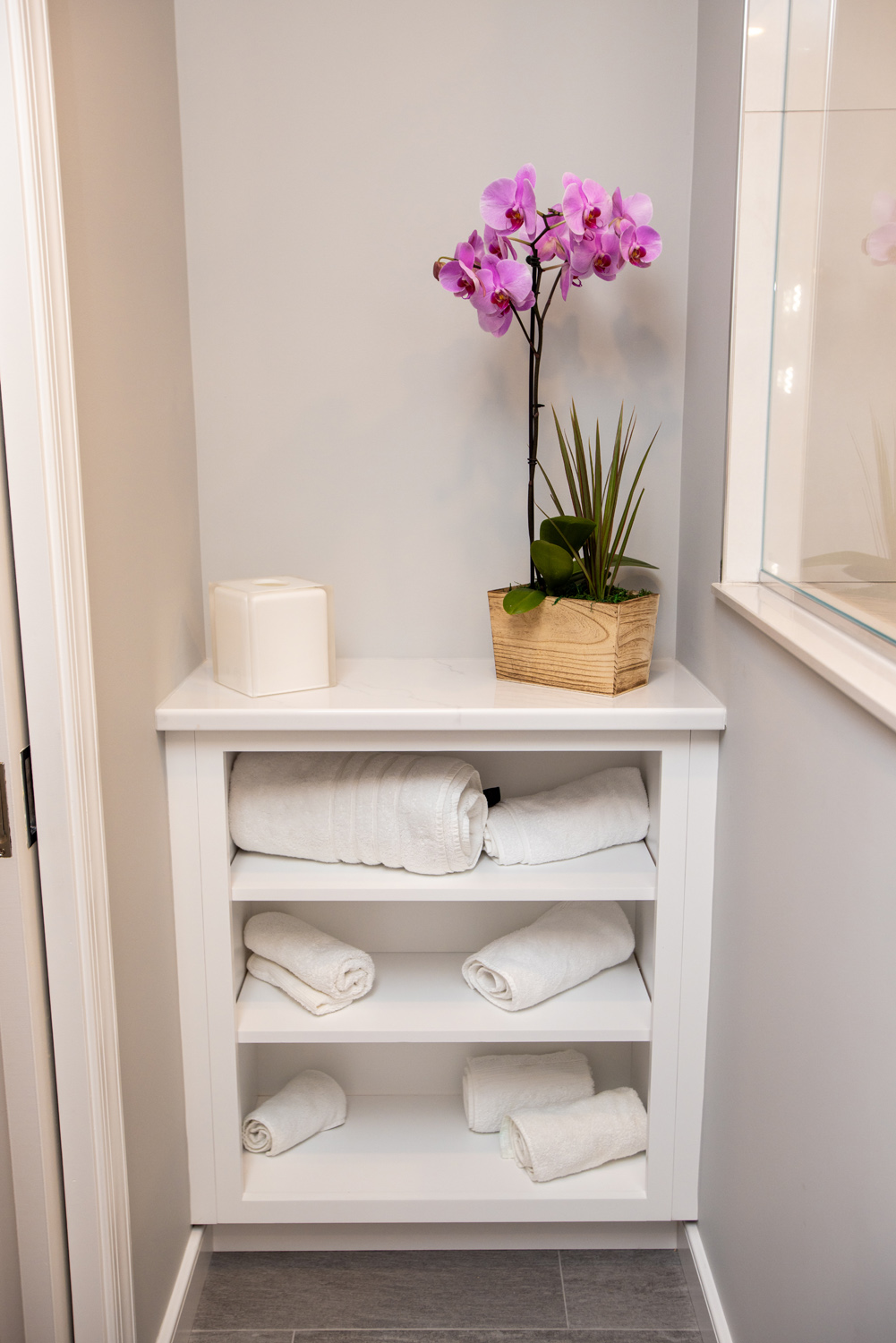 White shelving unit with neatly rolled white towels, a tissue box, and a potted orchid with pink flowers in a modern bathroom setting.