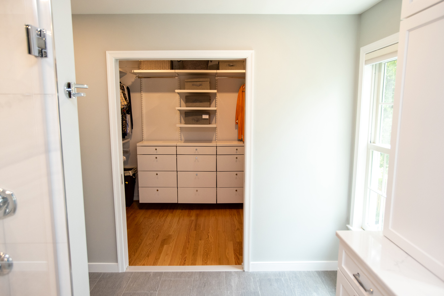 A walk-in closet with wooden flooring, white drawers, shelves, and hanging clothes, viewed from an adjoining room with tiled flooring and a window on the right.