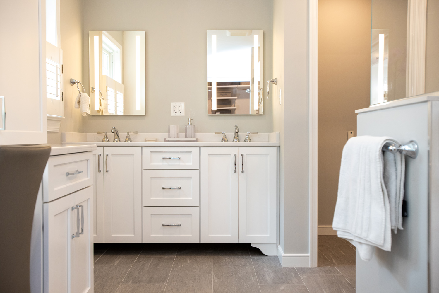 Bathroom with double sinks, white cabinets, two mirrors, and a towel hanging on a rack. The room has gray floor tiles and neutral wall colors.