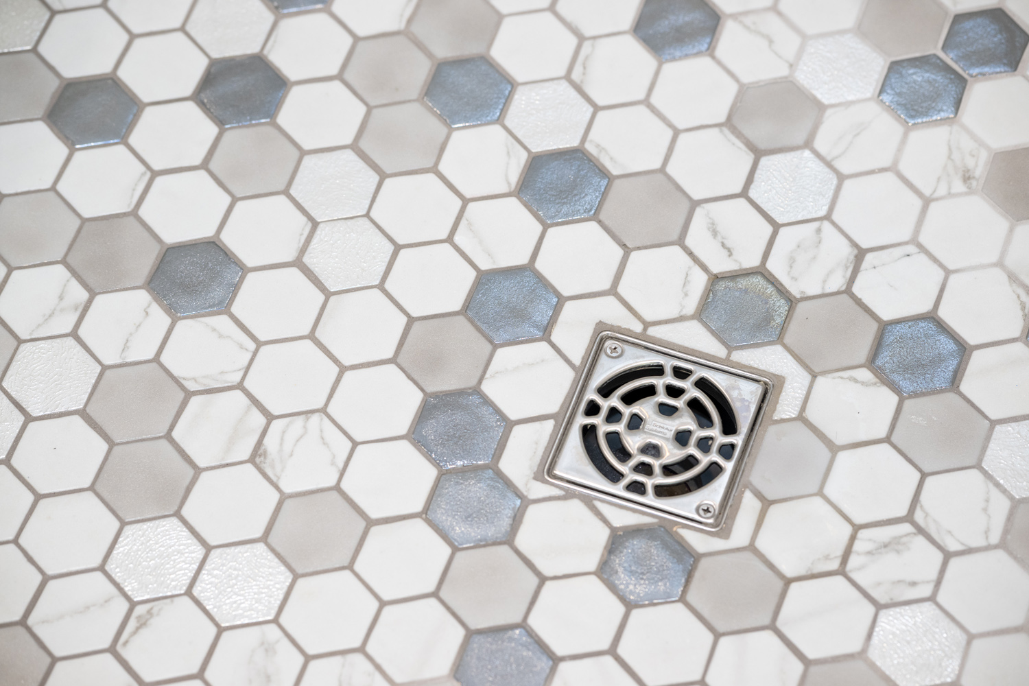 Hexagonal beige, white, and gray tiles arranged around a square metal shower drain cover on a bathroom floor.