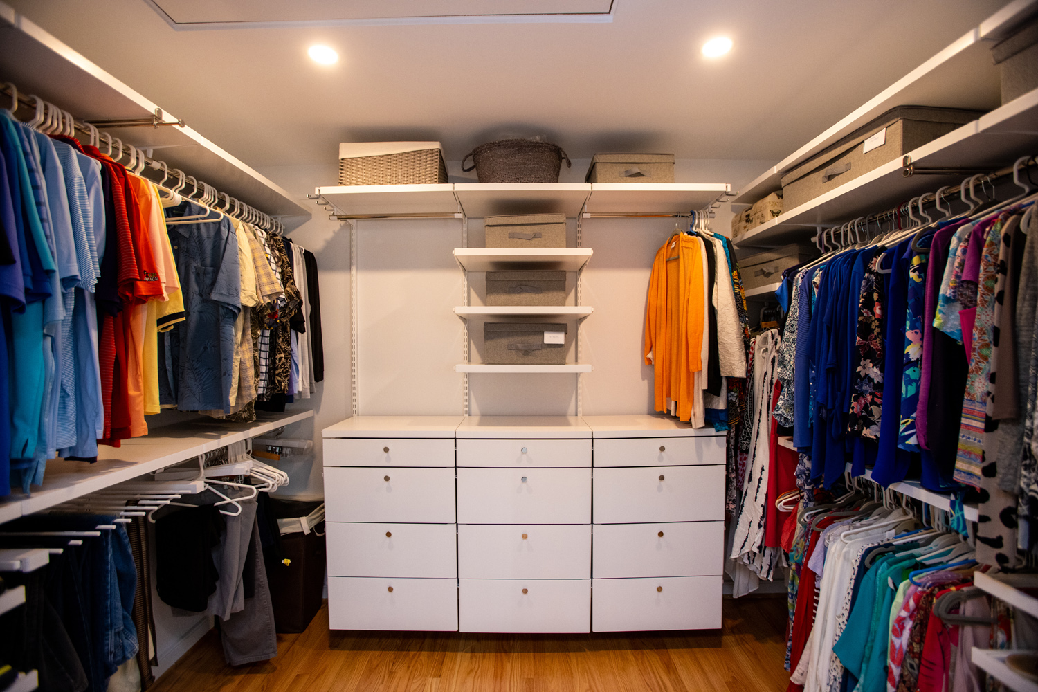A neatly organized walk-in closet with shelves, drawers, and hanging clothes on both sides, under warm ceiling lights.
