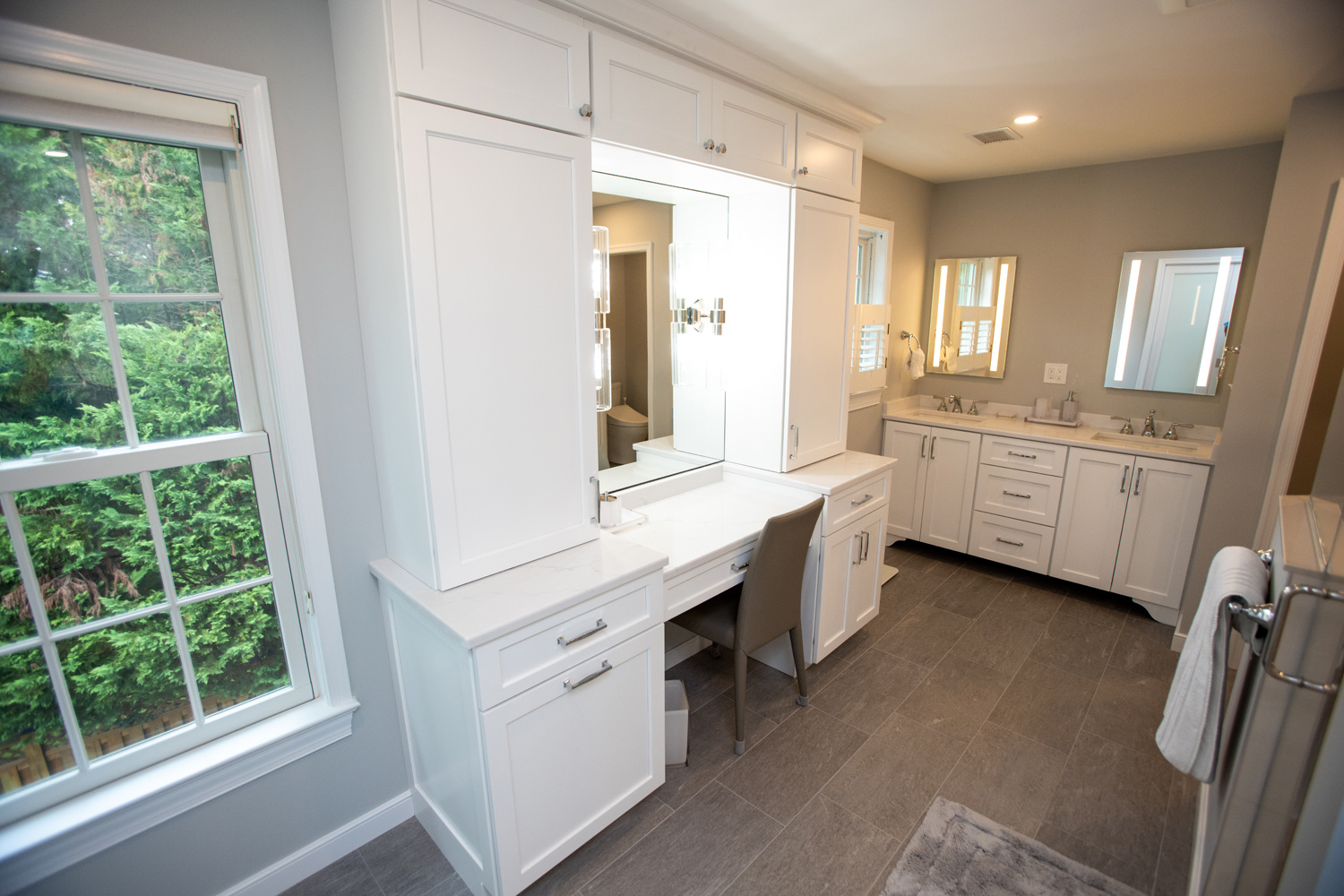 Modern bathroom with a large vanity area, chair, dual sinks, white cabinets, grey tile floor, and a window overlooking greenery.