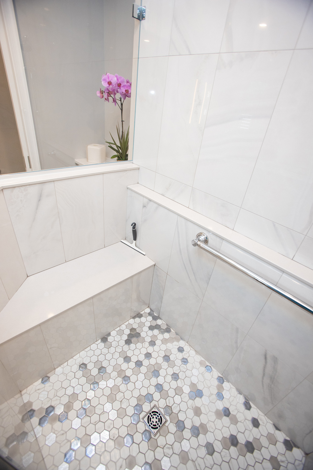White tiled shower with a corner bench, hexagonal tile floor, glass divider, grab bar, and a purple orchid on the ledge.